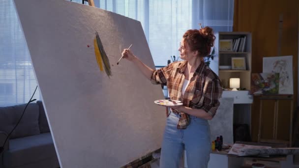 Sanatçı kız yavaş çekimde, gözlüklü genç kadın sehpada büyük bir tuval üzerinde fırçayla resim çiziyor. — Stok video