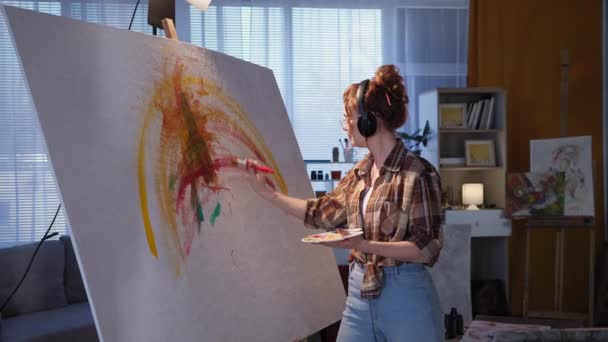 Dziewczyna w słuchawkach rysuje pędzlem na dużym płótnie przy użyciu jasnych kolorów, artysta wykorzystuje sztalugi podczas nowoczesnej kreatywności w studio — Wideo stockowe