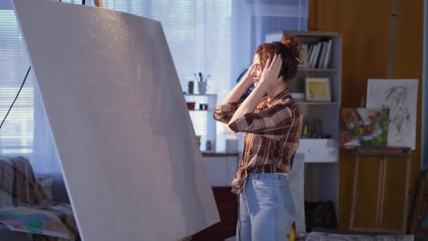 Umělec dívka nasadí sluchátka před kreslení procesu, zatímco stojí v blízkosti velkého stojanu, hudební a kreativní mladá žena v brýlích se štětcem na výtvarné dílně — Stock video