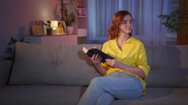Gözlüklü kadın elinde kitapla evde kitap okumaktan ve dinlenmekten hoşlanıyor.
