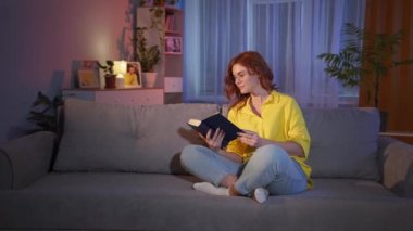 Gözlüklü kız elinde kitapla geceleri rahat bir odada kanepede oturup kitap okumaktan zevk alıyor.