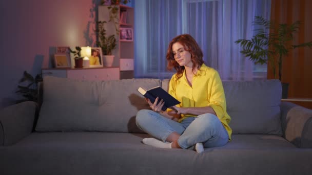 Wanita dengan kacamata beristirahat di kamar di malam hari dan membaca sebuah buku yang menarik di sofa dan menunjukkan ibu jari, memiliki ide — Stok Video