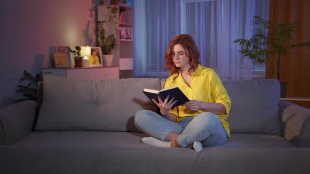 Wanita dengan kacamata menarik membaca buku di malam hari dan menunjukkan jempol sambil duduk di sofa di apartemen nyaman — Stok Video