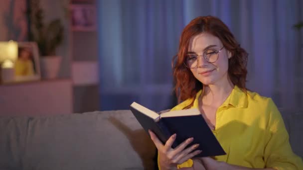 Femme avec des lunettes pour la vue profite du livre de lecture de détente du soir sur le canapé dans une chambre confortable, souriant et regardant la caméra — Video