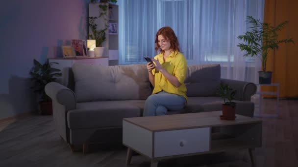 Femme en lunettes avec smartphone dans les mains montre pouce là-haut il ya une idée tout en étant assis sur le canapé à la maison, souriant et regardant la caméra — Video