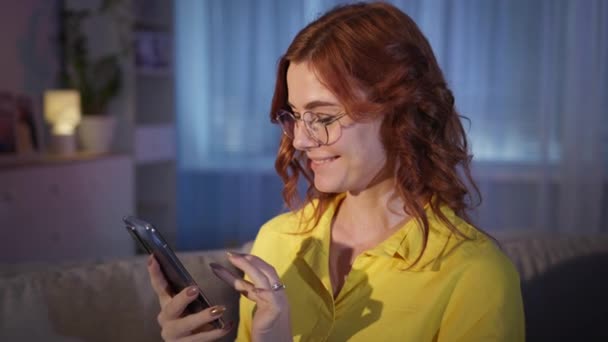 Kvinna med glasögon använder mobiltelefon och visa på produkter på Internet, ler och tittar på kameran när du sitter på soffan hemma på kvällen — Stockvideo