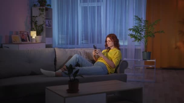 Donna con cellulare in mano si trova sul divano in soggiorno la sera a casa — Video Stock