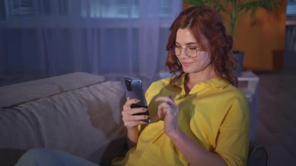 Kvinna med glasögon använder mobiltelefon och internet samtidigt som du kopplar av på soffan i vardagsrummet på kvällen — Stockvideo