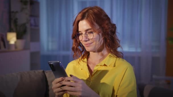 Frau mit Brille nutzt moderne Technik und genießt Handy, während sie abends zu Hause entspannt — Stockvideo