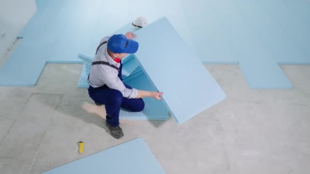 Homem em roupas de trabalho funciona como construtor no canteiro de obras e estabelece espuma de poliestireno expandida para isolamento térmico e colocação de laminado no chão, aponta para a câmera — Vídeo de Stock