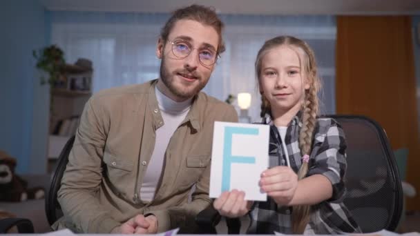 Rodinný portrét během online lekce, mladý otec s brýlemi pomáhá dceři dělat školní úkoly s kartami na fotoaparátu notebooku — Stock video