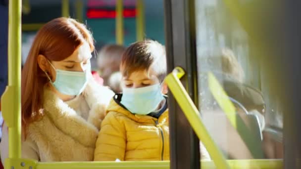 Jonge vrouw met zoon in de armen in medisch masker behandelt handen met antiseptische als gevolg van pandemieën tijdens het reizen met haar kind in de openbare bus — Stockvideo