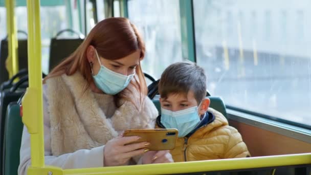 Kobieta z synem w maskach medycznych w celu ochrony przed zakażeniem koronawirusem grać w gry na smartfonie podczas podróży autobusem publicznym — Wideo stockowe