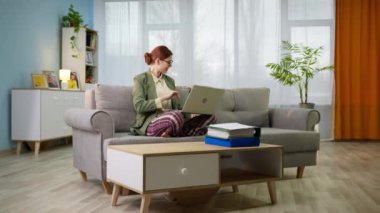 Kadın ofis çalışanı ceketli ve pijamalı bilgisayarda konuşurken evdeki koltukta uzaktan kumandayla çalışıyor.