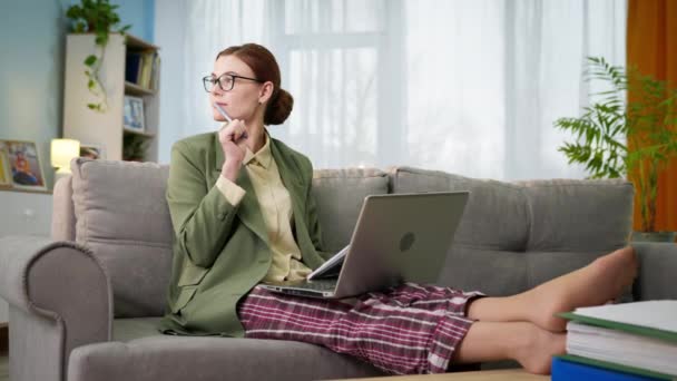 Mujer con chaqueta y pijama con portátil trabaja en casa de forma remota toma notas en documentos mientras está sentado en el sofá en la habitación — Vídeos de Stock