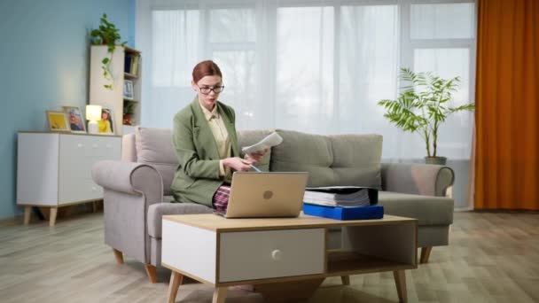 Kobieta w okularach z dokumentami w rękach działa online, rozmowy wideo na laptopie podczas siedzenia na kanapie w domu — Wideo stockowe