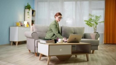 Ceketli ve pijamalı kadın ofis çalışanı evde uzaktan çalışıyor ve video çağrısı sırasında elinde bir bardak suyla odada kanepede oturuyor.