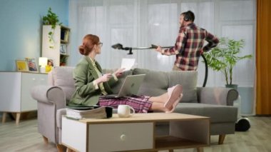 Ceketli ve pijama pantolonlu bir iş kadını laptoptaki video bağlantısıyla ofis müdürüyle konuşurken kulaklıklı bir adam odayı süpürüyor.