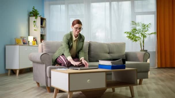 Kontorsarbetare i pyjamas och jacka arbetar på distans hemma sitter på soffan med en bärbar dator och dokument i händerna, frilans — Stockvideo