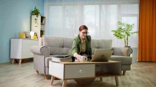 Mujer con chaqueta y pijama trabaja remotamente en casa y corre con una taza de agua en las manos a la computadora portátil durante la videollamada se sienta en el sofá en la habitación, el bloqueo — Vídeo de stock