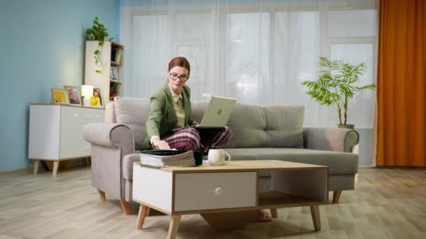 ジャケットとパジャマパンツの女性はノートパソコンで自宅でオンラインで作業し、リビングルームでソファに座っている間に文書を見て — ストック動画