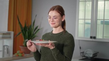 Kız tabağı mikroyeşilliklerle, domateslerle ve dolmalık biberlerle tutuyor, gözlüklü genç kadın salatadan hoşlanıyor sonra kameraya bakıyor ve mutfakta gülümsüyor.