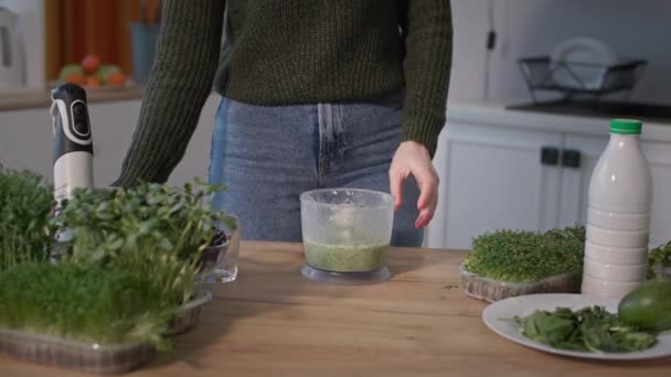 Menina corta microgreens com tesoura e, em seguida, mistura no liquidificador, jovem mulher derrama smoothie verde em um copo na cozinha — Vídeo de Stock