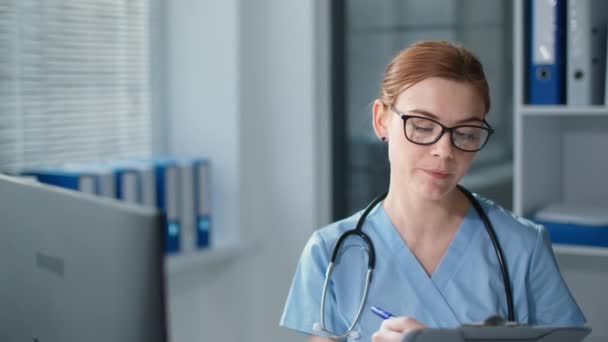 Médico fêmea atraente em uniforme azul com estetoscópio usa chamada de vídeo webcam para consulta on-line paciente, menina acenando mão e olha para a câmera com sorriso — Vídeo de Stock
