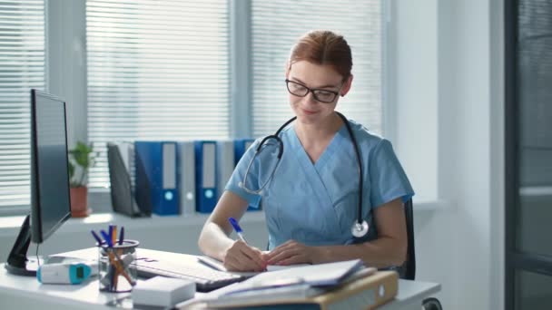 Retrato de la hermosa médico femenino vistiendo uniforme azul con estetoscopio, mujer sonriente en gafas escribe en el cuaderno y luego mira a la cámara en la oficina del hospital — Vídeos de Stock