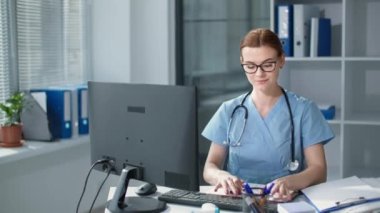 Hastane ofisindeki bilgisayarda çalışan kadın doktor asistanı. Gözlüklü mutlu doktor portresi. Masada steteskoplu mavi üniforma giyiyor.
