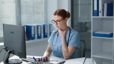 Hasta kadın doktor bilgisayarın başında çalışırken boğaz ağrısı hisseder. Gözlüklü doktor hastanede steteskoplu mavi üniforma giyer.