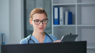 Üzerinde steteskop bulunan çevrimiçi doktor bilgisayar monitörünün önünde panoya yazıyor, gözlüklü genç kadın sağlık kliniğindeki hastasıyla konuşuyor.