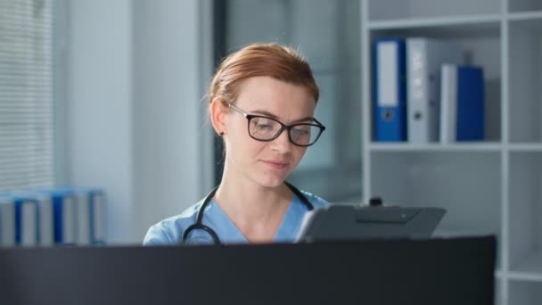 Portrait de belle thérapeute en uniforme bleu avec stéthoscope, souriante jeune femme dans les lunettes écrit dans le presse-papiers puis regarde la caméra au bureau de l'hôpital — Video