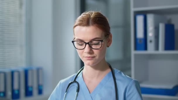 Portret lekarza w okularach podczas pracy w szpitalu, młoda terapeutka pisząca na klawiaturze komputera, a następnie patrząca w kamerę z uśmiechem z bliska — Wideo stockowe