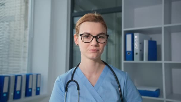 Médecine, femme médecin en lunettes portant un uniforme bleu avec stéthoscope parlant en ligne avec le patient et regardant la caméra avec presse-papiers à la main — Video