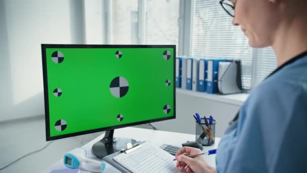 Arzt berät Patient online über Computer mit grünem Bildschirm, Arzt in blauer Uniform spricht und schreibt in Klemmbrett am Arbeitsplatz im Krankenhaus — Stockvideo