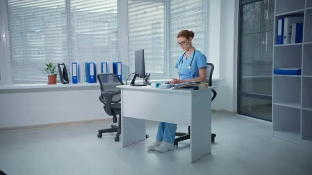 Εργατικός οικογενειακός γιατρός εργάζεται σε ένα γραφείο νοσοκομείου, γυναίκες θεραπευτές τύποι στο πληκτρολόγιο υπολογιστών αισθάνεται έπειτα έναν πονοκέφαλο ενώ κάθεται σε ένα τραπέζι με τα έγγραφα — Αρχείο Βίντεο