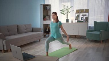 Eşofmanlı genç kadın, yoga öğretmeniyle yoga minderi üzerinde video bağlantısı ve yaşam tarzı üzerinden fiziksel egzersiz yapıyor.