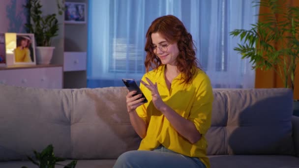 Mulher com óculos usa telefone celular para fazer compras on-line, sentado no sofá durante o relaxamento à noite na sala de estar — Vídeo de Stock
