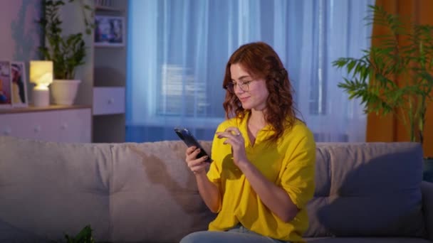 Lány szemüvegben, mobiltelefon a kezében, mosolygós és a kamerába néz, miközben ül a nappaliban este — Stock videók