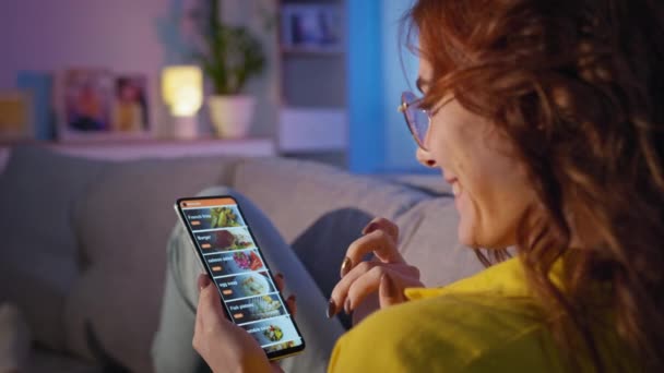 Молодая женщина с мобильным телефоном в руках делает покупки в Интернете, выбирая фаст-фуд на сайте ресторана или кафе, сидя на диване — стоковое видео