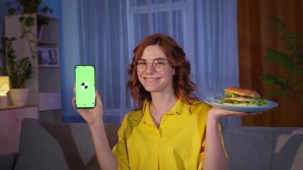 Encantadora mujer sonriente con hamburguesa y teléfono inteligente con tecla de croma verde en la pantalla se sienta en el sofá en la habitación por la noche sonriendo y mirando a la cámara — Vídeos de Stock