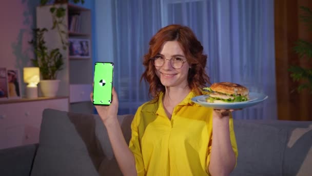 Donna in occhiali con hamburger e telefono cellulare con chiave cromatica verde in mano sorride e guarda alla telecamera durante uno spuntino serale a casa — Video Stock