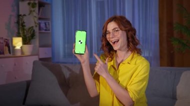 Gözlüklü kız evde kanepede dinlenirken yeşil krom anahtarlı cep telefonunu ekrandan ekrana gösteriyor.