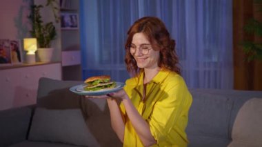 Aç, neşeli bir kadın, elinde hamburgerle akşam yemeğinde evde dinleniyor.