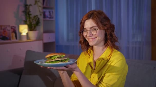 Kobieta w okularach z talerzem hamburgera w dłoniach raduje się z fast foodów siedzących wieczorem na kanapie w domu — Wideo stockowe