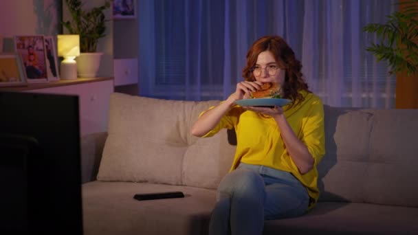 Vrouw eten fast food hamburger in de voorkant van TV terwijl zitten op de bank thuis in de avond, thuis blijven — Stockvideo