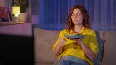 Gözlüklü yetişkin bir kız, akşam karantinada hamburger yer ve komik bir program izler.