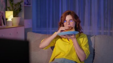 Gözlüklü neşeli kız TV 'de komik programlar izleyerek gülüyor, geceleri kanepede otururken fast food yiyor, karantina altında.