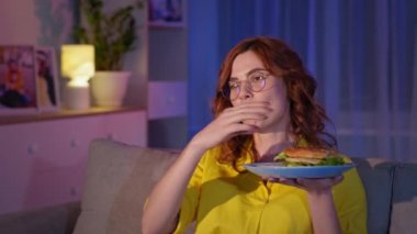 Ev hanımı hamburgerini yerken kanepede eğlenen komik bir TV programı izliyor.